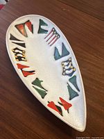 Full view of elongated oval shaped ceramic bowl with cream textured glaze and colorful abstract geometric decorations around rim.