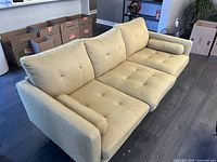 Full view of yellow sofa couch seen from an angle, showing three seat cushions, back cushions, and one bolster pillow on the left side. Floor and some unpacked boxes in background.