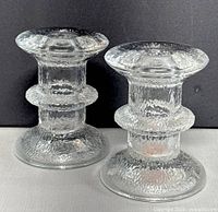 Front view of a pair of textured clear glass candle holders showing the flared top, ring detail at midsection, and broad base on a neutral background.