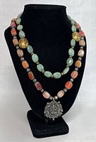 Front view of two stacked necklaces on black display: top necklace with light green jade-like beads and gold accents; bottom necklace with multi-colored sunstone-like beads, silver beads and detailed pendant.