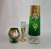 All three glassware pieces shown frontally to capture size and details. The tallest is a green and gold footed vase, accompanied by a green and gold goblet and a blue and gold flute glass with painted floral patterns.