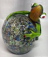 Art glass frog climbing on speckled spherical base, detailed view of frog's face and front limbs.
