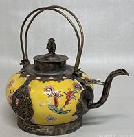 Front view of the teapot showing yellow porcelain body with floral designs and silver brass dragon accents, lid with monkey figurine, curved double handle, and spout.