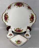 Front view of large cake plate, pair of salt and pepper shakers, and small square dish showing Royal Albert Old Country Roses pattern and gold accents.