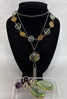 Front view of the necklace on a black bust showing art glass beads and large pendant with tassel, earrings in front.