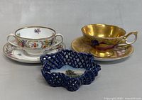 Photo showing the vintage items including a fruit patterned teacup with saucer, cobalt blue basket, and a white floral double handled teacup with saucer.