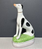 Front-side view of ceramic dog figurine showing the sitting posture, black spots, and gold collar on a green grassy base.