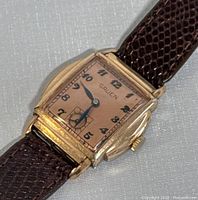 Front view of watch face showing pink dial, blue leaf hands and Arabic numerals