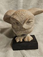 Front view of beige owl sculpture showing detailed head and eyes mounted on a black base on soft backdrop.