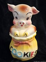 Front and angled view of bisque pottery pig cookie jar holding a yellow sack labeled 'COOKIES'.