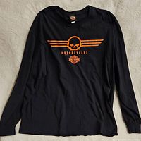 Full view of black long sleeve Harley Davidson shirt with orange skull and wing logo and text 'Motorcycles' with Harley Davidson shield logo underneath.