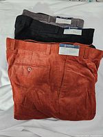 Three pairs of men's pants folded, showing colors: rust corduroy, black corduroy, gray dress pants.