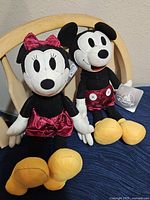 Photo showing Mickey Mouse and Minnie Mouse plush toys sitting on a chair; both have red clothing with yellow shoes and black and white features.
