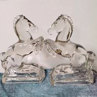 Front view of both clear glass horse figurines side by side on blue fabric, showing raised front legs and overall shape.