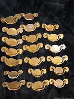 Overview of 18 vintage brass drawer pulls laid out on black background, showing front face and detailing of Chippendale bail handle design.