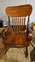 Full view of a vintage wooden rocking chair with a curved backrest, armrests, and contoured seat placed on floor tiles in front of window blinds.