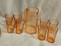 Full set of one pink swirl glass pitcher surrounded by six matching glasses on a white fabric background.