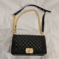 Full view of the new black quilted Emperia purse lying on white fabric, showing gold chain and black leather strap with tagged label attached.