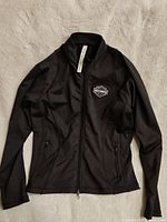 Front view of black women's zip-up jacket laid flat showing full design, zippers, and Harley Davidson patch