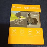 Front of new box showing the Yada 720P Roadcam dash camera with visible lens and suction cup mount behind plastic window. Box highlights HD 720P, 2 inch screen, G-Sensor Technology, and Motion Detection icons.