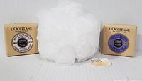 Two L'OCCITANE soap bars and a white bath pouf displayed on a white textured surface.
