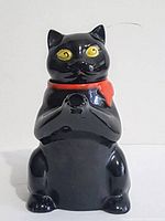 Front view of black ceramic cat-shaped tea pot sitting upright with yellow eyes, red scarf, and spout at front.