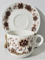 Side front view of the ceramic cup showing detailed brown floral patterns with the saucer behind it
