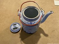 Side angle view of white ceramic tea pot with blue decorative design, orange handle, and matching lid beside it on the surface.