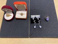 Full view of all jewelry items displayed on black foam: two rings in red and orange boxes, pair of floral silver-tone earrings on black card, silver-tone ring with lavender stone.