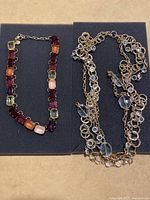 Two costume jewelry necklaces displayed on black foam; left necklace with multicolored square stones, right necklace gold-tone with clear stones.