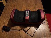 Foot massager and warmer placed on a wooden surface showing the top side with control panel and attached power cord.