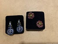 Both pairs of earrings shown on black display pads: blue geometric drop earrings on the left, pink floral stud earrings on the right.
