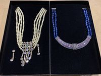 Two necklaces displayed side by side in black velvet box; one cream pearl style with crystal accents and matching earrings, the other blue beaded with purple stones.