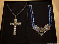 Two necklaces in black display box: one a cross pendant necklace with clear rhinestones on a black cord; the other a two strand blue beaded necklace with floral clusters of pink, purple, and blue stones