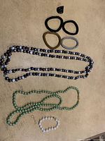 Multiple bracelets of various colors including black, green, gold, silver, and clear beads, and two long beaded necklaces in dark metallic and green beads.