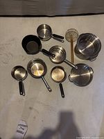 Top-down view of various kitchen pots and utensils arranged on a floor, showing different sizes and handle types.
