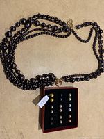 Black multi-strand beaded necklace displayed flat showing the gold-tone KJL clasp alongside the set of assorted stud earrings in a box.