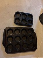 Two black metal muffin tins placed on a surface, one with 6 muffin cavities and the other with 12.