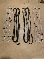 Four long beaded necklaces laid flat on a beige surface: two white strand necklaces with multicolored spherical accent beads, and two black strand necklaces with multicolored spherical accent beads.