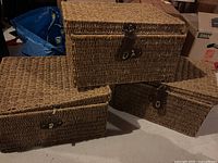Three square wicker chests stacked, showing woven texture and metal buckles, with some buckles damaged.