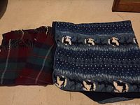 Photo showing two blankets: a folded plaid flannel blanket in dark red and green tones and a folded fleece blanket with blue background featuring white and black reindeer and snowflake patterns.