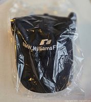 Front view of black BMW Williams F1 cap still sealed in clear plastic showing embroidered team logo on front.