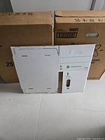 Two closed cardboard shipping cases labeled for 25 boxes each with folded flat white cardboard box samples leaning against them showing the Grand & Toy record storage boxes.