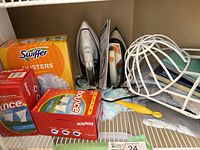Ironing board and cleaning supplies including Bounce dryer sheets, Swiffer dusters, irons, and baseball hat dishwasher bracket on wire shelving.
