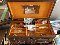 Opened brown vintage-style jewelry box with orange interior, showing assorted costume jewelry pieces including rings, chains, brooches, and small pins.