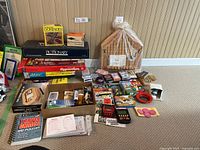Wide shot showing multiple board games stacked including Scrabble, Pictionary, Aggravation, dominoes, card games, and boxed sets with a wooden card holder box.