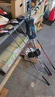 Four golf clubs with head covers leaning against a workbench, Daiwa ball picker, and open box of golf balls visible