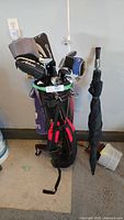 Full set of golf clubs in a black and red golf bag standing upright next to a folded black umbrella.