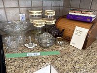 Photo of glass punch bowl, clear glass cups, Pyrex containers with lids with floral decals, a dark brown teapot, metal tea ball strainer on dish, bread box, and recipe books on kitchen counter.