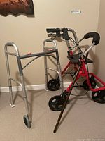 Metal two-wheeled walker next to red four-wheeled walker and wooden cane